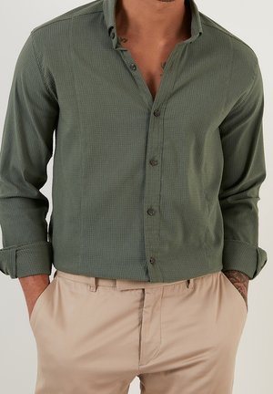 Homme portant une chemise boutonnée vert olive aux manches retroussées et un pantalon beige, les mains dans les poches, un tatouage visible sur le poignet droit.
