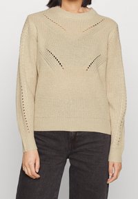 Beige stickad tröja med ribbade muddar och nederkant. Med diagonala utskärningar på framsidan och subtila öppna stickade detaljer på ärmarna.