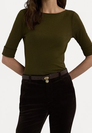 Woman wearing olive green fitted top with elbow-length sleeves and black corduroy pants with a dark belt featuring a gold buckle charm.