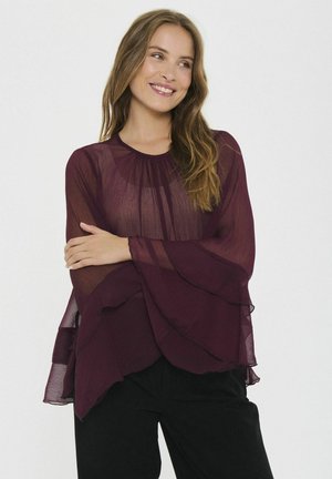 Blusa trasparente bordeaux con maniche fluide e arricciate e scollatura rotonda, abbinata a pantaloni neri. Tessuto leggero che cade morbido.
