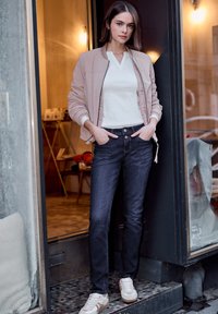 Jeune femme aux cheveux bruns courts se tenant dans l'encadrement d'une porte, portant une veste bomber rose pâle, une chemise blanche, un jean foncé et des baskets blanches.