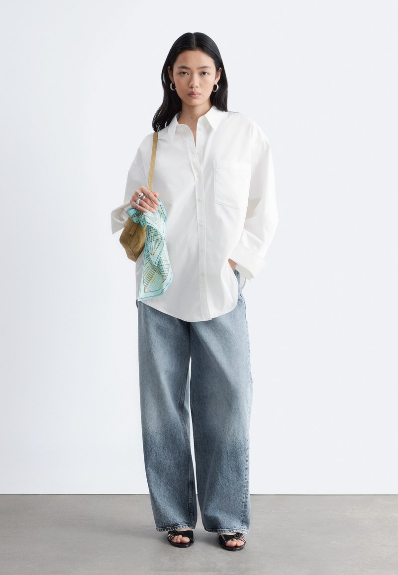 Femme portant une chemise blanche à boutons oversize, un jean à jambes larges, des sandales noires ouvertes et portant un sac beige avec une écharpe bleue à motifs.