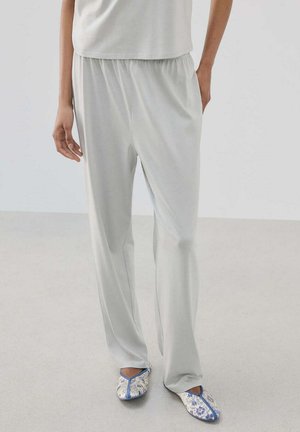 Personne portant un pantalon ample gris clair et des chaussons blancs avec des motifs floraux bleus, debout sur un sol clair contre un mur uni.
