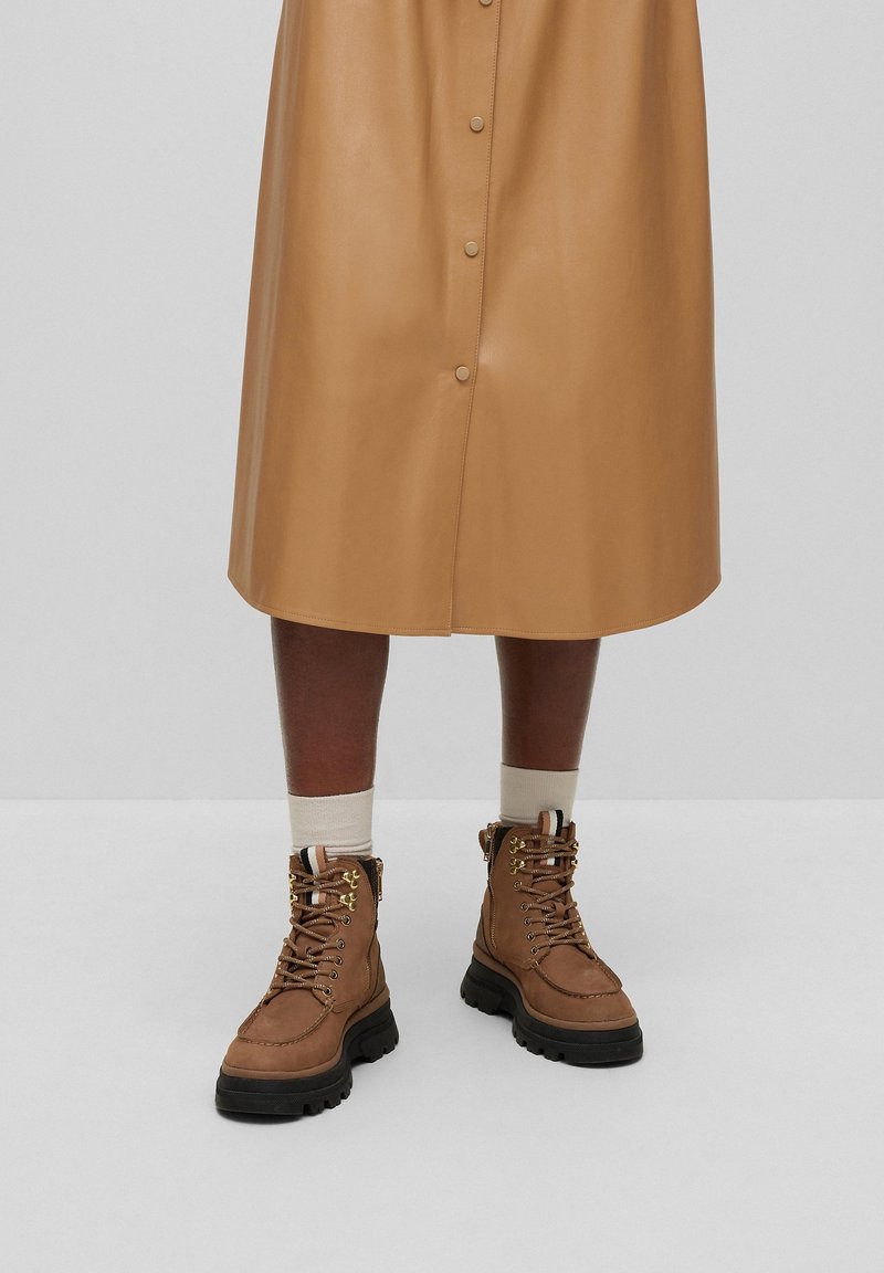 Botas marrones con cordones, suela robusta, parte superior de cuero y herrajes dorados. Llevadas con calcetines de color claro y una falda abotonada en tono beige.