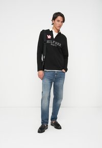 Jaunietis, ģērbts melnā Hilfiger Racing džemperī, zilos džinsos, melnos fraku kurpēs un ar melnu pleca somu, stāv uz baltas fona.