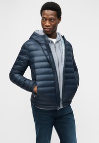 Άνδρας που φοράει σκούρο μπλε φουσκωτό μπουφάν πάνω από ανοιχτό γκρι φούτερ με κουκούλα και τζιν, στεκόμενος με τα χέρια στις τσέπες του μπουφάν, κοιτάζει προς τα πλάγια.