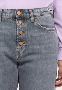 Jeans de mezclilla gris de talle alto con cinco botones, herrajes de latón y detalles de dobladillo deshilachado. Presentan bolsillos clásicos y una textura sutil.