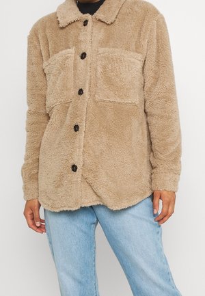 Beige fleecetrøje med knapper og to brystlommer, båret over sort top, kombineret med lyseblå denimjeans.