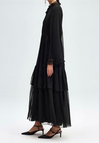 Robe longue noire à volants avec un tissu transparent, accents froncés aux manches et au col, associée à des escarpins noirs à bout pointu avec des brides de cheville.