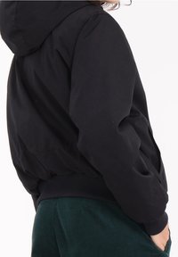 Giacca nera con cappuccio, dal fit rilassato, con polsini e orlo a coste, realizzata in tessuto liscio, mostrata di lato su uno sfondo neutro.