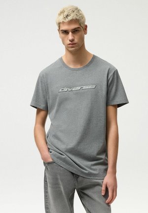 Młody mężczyzna o krótkich blond włosach, ubrany w szarą koszulkę z logo "diverse" oraz pasujące szare jeansy, stojący z jedną ręką w kieszeni.