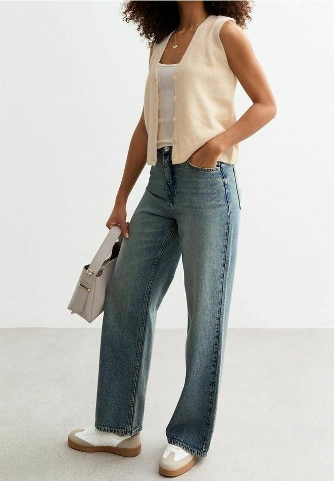 Kremowa dzianinowa kamizelka, biały top na ramiączkach, luźne niebieskie dżinsy, białe buty w stylu trampek z brązowymi podeszwami, trzymająca małą beżową torebkę.