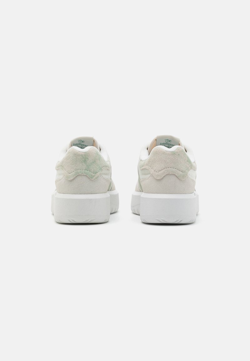 Un paio di sneaker in suede beige chiaro con suole bianche spesse viste da dietro su uno sfondo bianco uniforme.