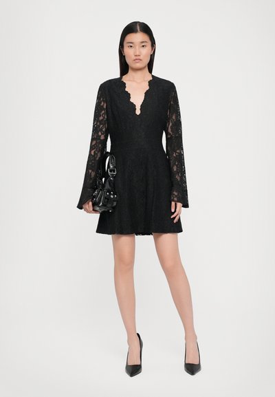 Black lace dress with long bell sleeves, deep V-neckline, fitted waist, flared skirt. Model holds a small black handbag.