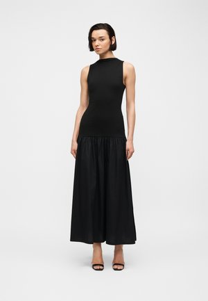 Kvinde med kort mørkt hår iført en ærmeløs, tætsiddende sort top med et langt, samlet sort skørt og sorte åbne hæle, på hvid baggrund.