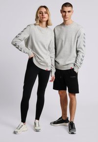 Grijze sweatshirt met contrasterende chevronpatronen op de mouwen, gecombineerd met zwarte leggings en shorts; beiden dragen sportieve sneakers.