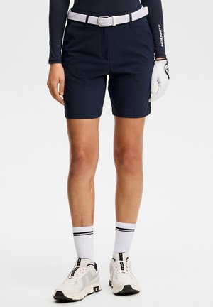 Person iført marineblå golfshorts med hvidt bælte, hvid golfhandske, hvide sneakers og hvide sokker med sorte striber.