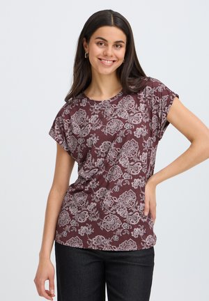 Jeune femme aux longs cheveux foncés sourit, portant un haut bordeaux à manches courtes avec des motifs floraux blancs et un pantalon noir.