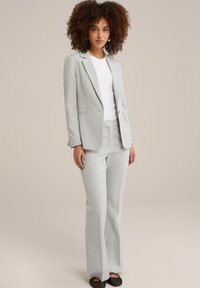 Traje ajustado de color gris claro con una chaqueta entallada y pantalones acampanados, confeccionado en una tela suave. Camisa blanca debajo y zapatos negros tipo slip-on.