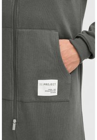 Dunkelgrauer Reißverschluss-Hoodie mit einer Kängurutasche, gerippten Bündchen und einem weißen Label, das "11 PROJECT" und Markendetails zeigt. Weicher Materialtextur.