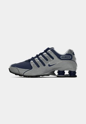 Nike Shox tekači čevlji z modrim zgornjim delom in sivimi poudarki, teksturiranimi materiali ter edinstvenim podplatom s spominskimi stebrički v peti.