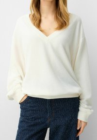 Biały sweter z dekoltem w serek wykonany z miękkiej, strukturalnej tkaniny. Luźny fason z szerokimi rękawami i ściągaczami przy mankietach. Połączony z ciemnymi, teksturowanymi dżinsami.