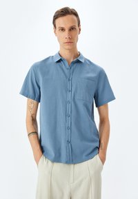 Korte mouwen, lichtblauw shirt gemaakt van een zachte stof, met een knoopsluiting aan de voorkant en een borstzak. Glad textuur en comfortabele pasvorm.
