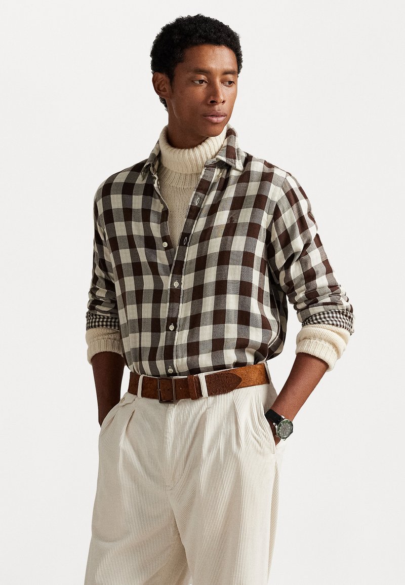Camisa de franela a cuadros marrón y crema, llevada sobre un cuello de tortuga de punto crema, combinada con pantalones crema con pliegues y un cinturón marrón.