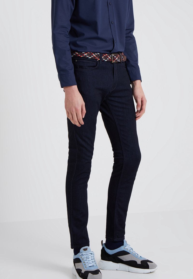Homme portant un jean slim foncé, une chemise bleu marine boutonnée, une ceinture à motifs, et des baskets bleu clair et noir sur un fond uni.