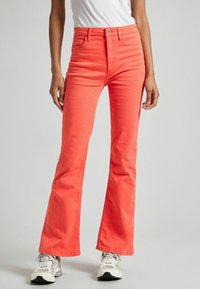Jeans acampanados en coral brillante, hechos de denim. Presentan un diseño clásico de cinco bolsillos y un cierre de botón. Llevados con zapatillas blancas.