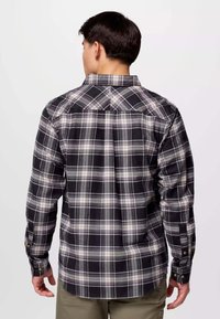 Uomo di spalle che indossa una camicia a maniche lunghe a quadri neri e grigi con polsini abbottonati e pantaloni verde oliva, su uno sfondo neutro.
