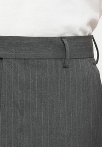 Detailní záběr na šedé proužkované kalhoty v oblasti pasu s poutky na opasek, nošené přes bílou košili.