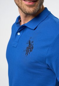 Muž se usmívá a má na sobě modré polo tričko s černě vyšitým logem hráče polo a koně na levé hrudi.