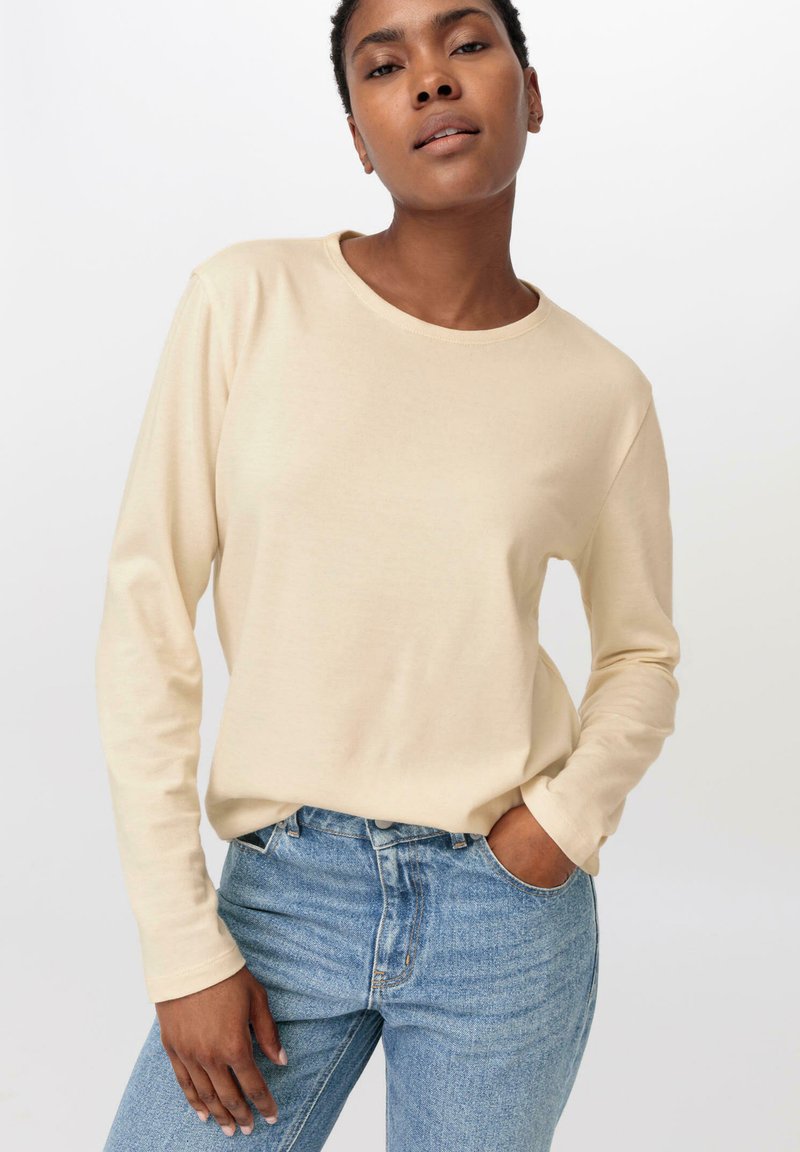Langärmliges, beiges T-Shirt aus weichem Stoff, mit rundem Ausschnitt und lässiger Passform, kombiniert mit hellblauen Jeans.