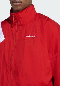 Červen á nylonová bunda s vysokým golierom, zapínaním na zips a bielymi detailmi. Obsahuje vyšívané logo Adidas na hrudi s textúrovaným povrchom.