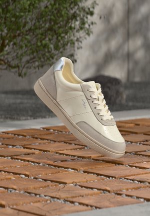 Beige og cremefarvet sneaker med sølv hældetalje, snørebånd bundet, placeret over en flisebelagt træs overflade med sløret grønt område og væg i baggrunden.