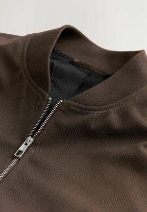 Chaqueta marrón con cremallera y cuello acanalado, que presenta una textura suave y cremallera plateada. El interior tiene un forro de malla.