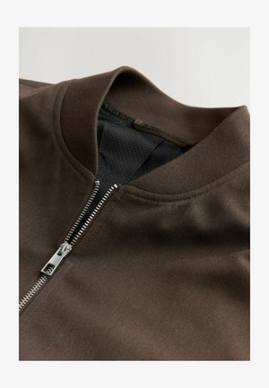 Chaqueta marrón con cremallera y cuello acanalado, que presenta una textura suave y cremallera plateada. El interior tiene un forro de malla.