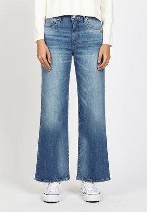Woman wearing light blue wide-leg jeans, white long-sleeve top, and white sneakers, standing against a plain background.