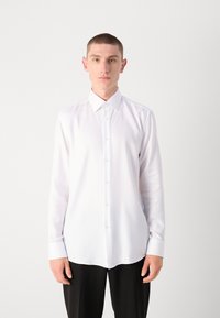 Chemise blanche à manches longues en tissu texturé, avec un boutonnage sur le devant et un col classique ; associée à un pantalon noir.