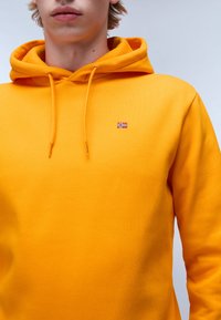 Gele katoenen hoodie met een voorzak, voorzien van een klein geborduurd vlagdetail op de borst en verstelbare trekkoorden bij de capuchon.