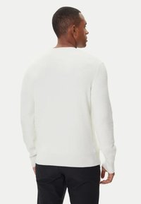Uomo di spalle che indossa un maglione bianco a maniche lunghe con texture e pantaloni neri, davanti a uno sfondo chiaro e uniforme.
