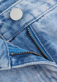 Gros plan d'un jean en denim bleu partiellement déboutonné, montrant une fermeture éclair en métal et un bouton métallique à logo.