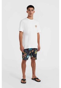 Witte katoenen t-shirt met een kleine kleurrijke graphic, gecombineerd met zwarte zwemshorts met een levendige tropische print in verschillende kleuren. Slippers aan de voeten.