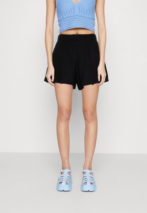 Femme portant un short noir taille haute, un crop top tricoté bleu clair et des sandales bleu clair à larges brides, debout devant un fond uni.