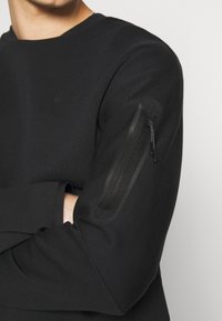 Musta collegepaita pyöreällä kaula-aukolla, jossa on vetoketjullinen tasku vasemmassa hiharesassa ja sileä, teksturoitu kangas. Hienovarainen logoyksityiskohta.