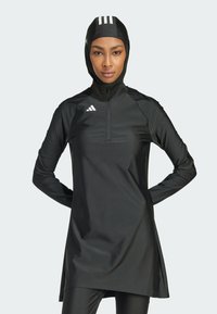 Robe de sport noire avec un col montant, des manches longues et une fermeture éclair. Présente des rayures blanches sur la capuche et le logo Adidas sur la poitrine.