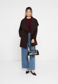 Abrigo de lana marrón con un collar ancho, combinado con un suéter burdeos, jeans de pierna ancha de color azul claro y tacones negros de puntera afilada, sosteniendo un bolso negro.