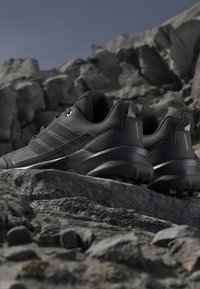 Scarpe da corsa trail nere per esterni su terreno roccioso con suole robuste e linguette sotto un cielo sereno.