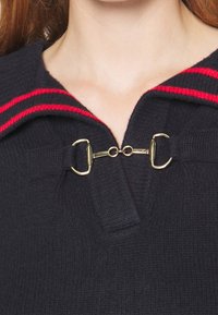 Marinblå stickad tröja med en vid krage med röda ränder, fäst med guldfärgad metall i ett dekorativt spänne vid halsringningen.